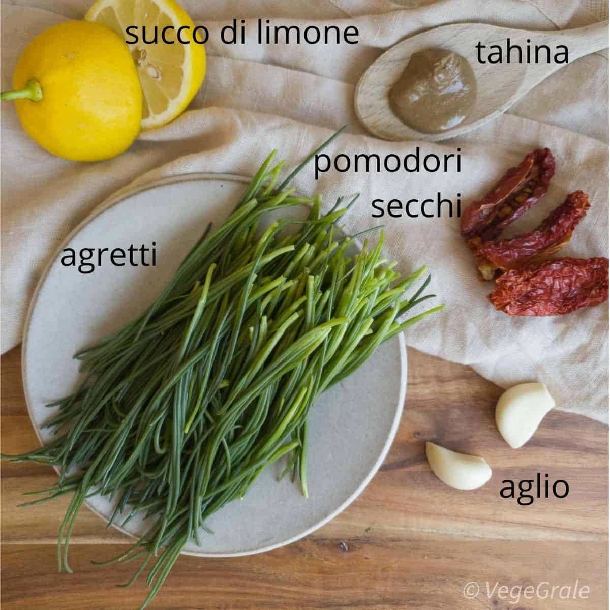 Si vedono tutti gli ingredienti necessari con le scritte affianco: agretti, aglio, pomodori secchi, tahina e limone.