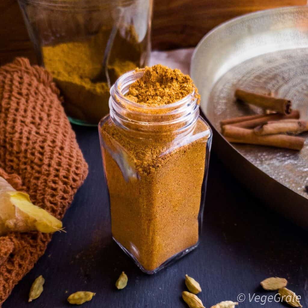 Polvere marrone in contenitore di spezie in vetro aperto. Intorno sono poggiati alcune spezie integrali, come cardamomo, stecche di cannella e zenzero fresco.