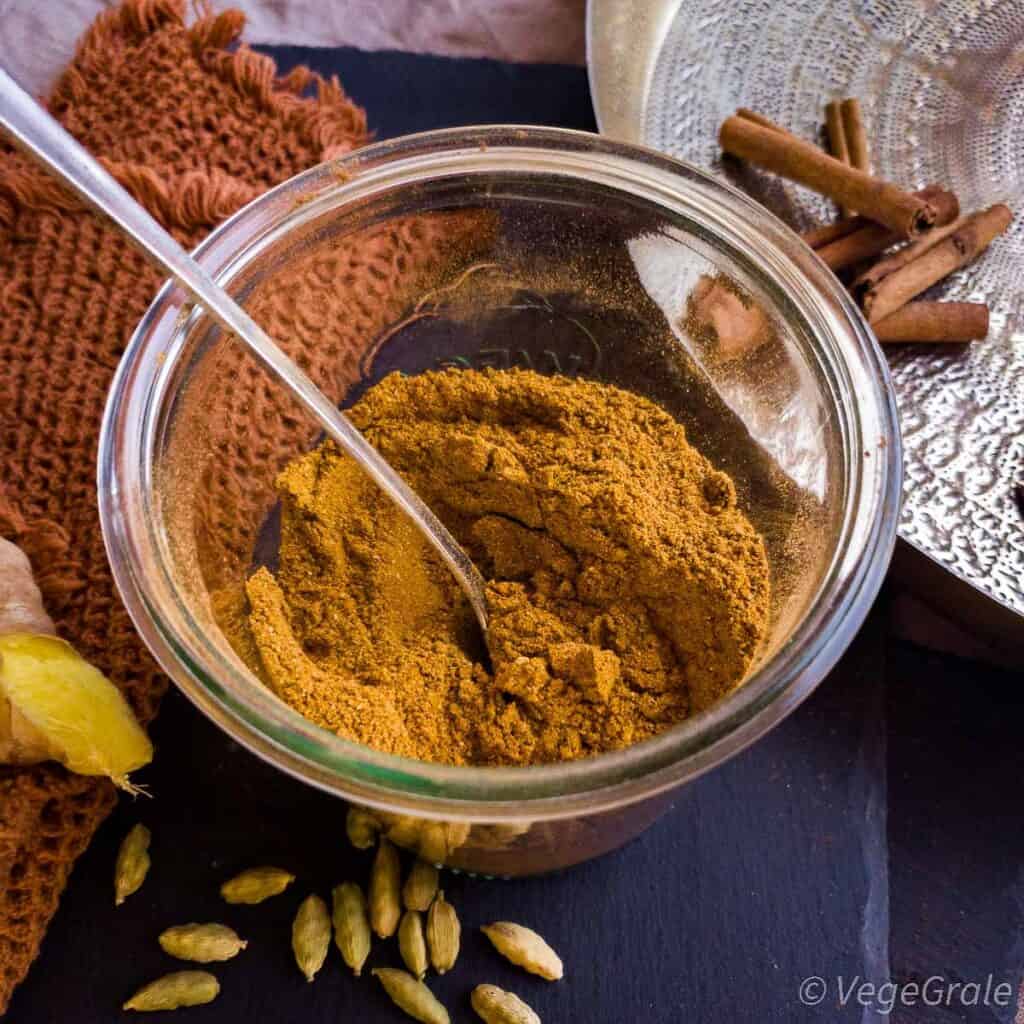 Polvere marrone in contenitore grande di vetro. Sul tavolo alcune spezie integrali, come cardamomo, stecche di cannella e zenzero fresco.
