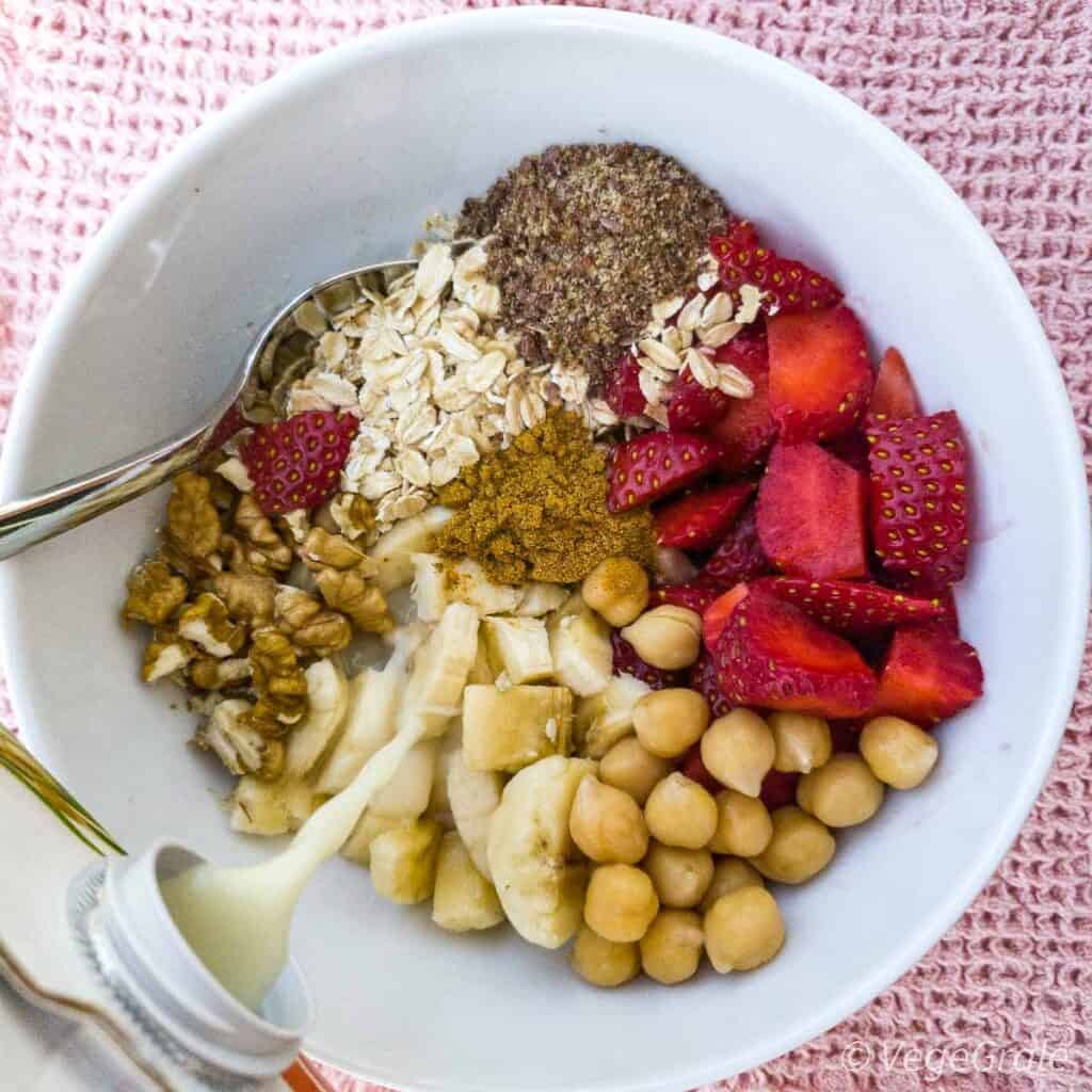 Ciotola da colazione con dentro fiocchi di avena, semi di lino macinati, fragole, ceci cotti, banane e noci. Viene versato del latte vegetale.