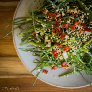 Insalata di agretti crudi condito con pomodori secchi, aglio e sesamo su piatto su tavolo di legno.