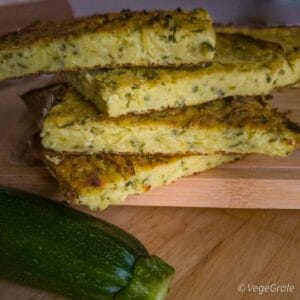 Pila di fette di farinata alle zucchine su tagliere, d'avanti appoggiata una zucchina cruda.