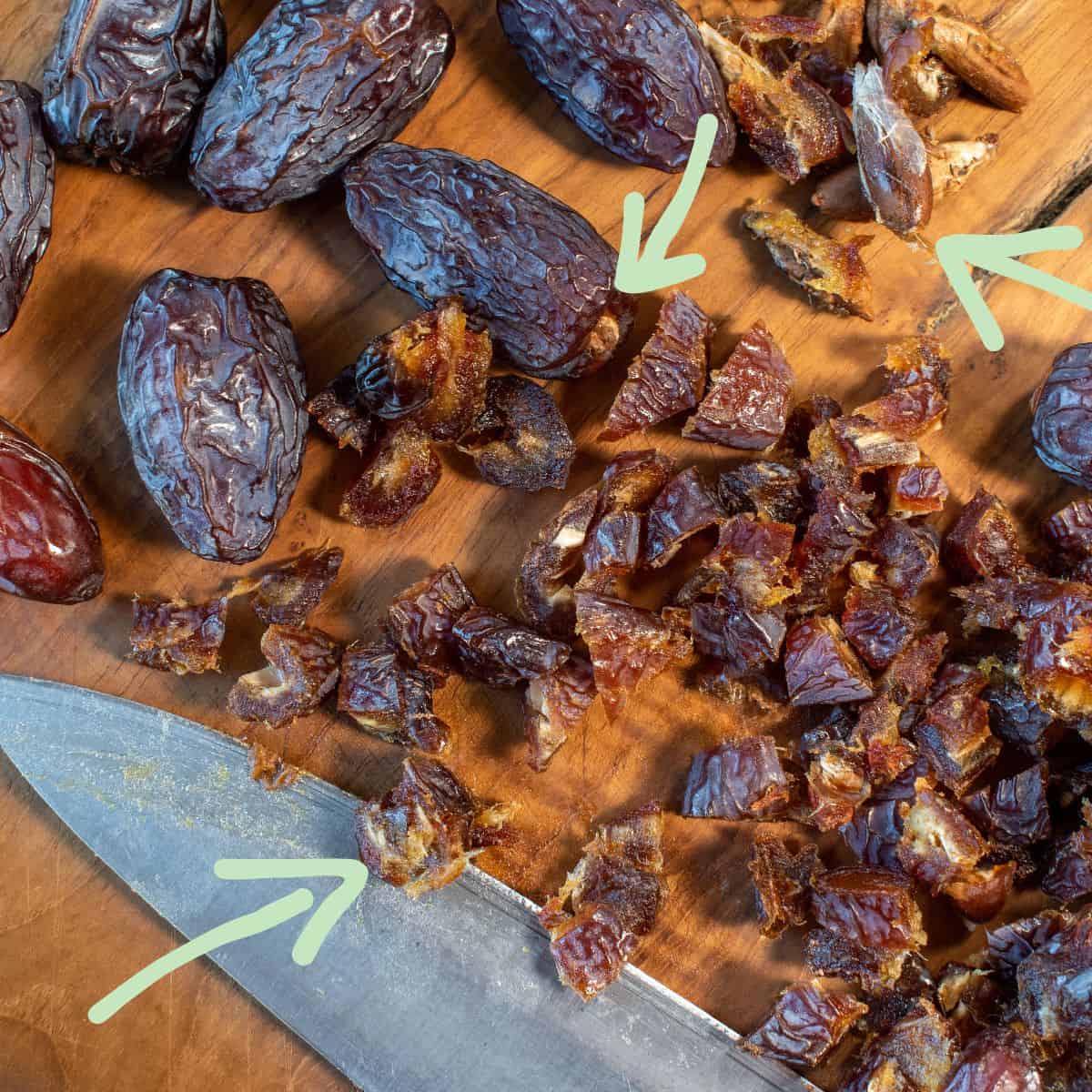 Tagliere con tanti datteri sopra. Alcuni interi, alcuni snocciolati e sminuzzati a pezzettini di mezzo centimetro circa. Si vede anche la punta di un coltello e una piccola pila di semi. Una freccia indica la parte dura sull'estremità del dattero intero, un'altra punta su un nocciolo estratto e un'altra freccia ancora su un pezzo di dattero tagliato.